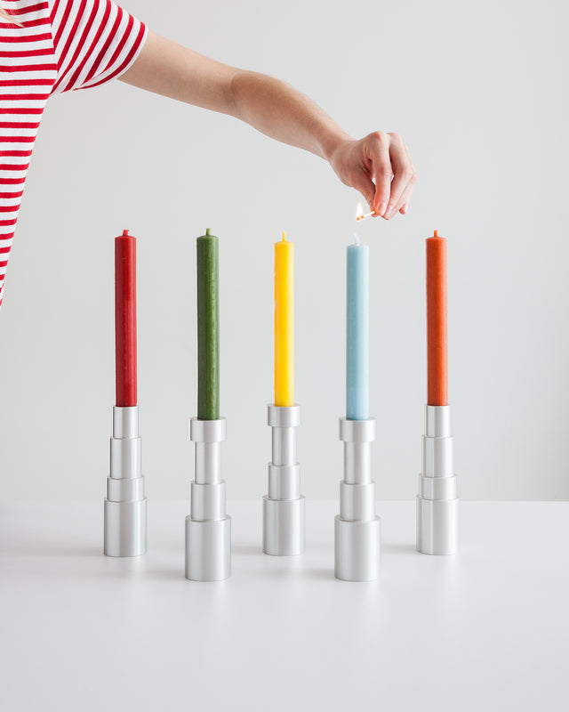 A row of five aluminum candle holders with colorful candles, displayed on a white surface against a white wooden background.